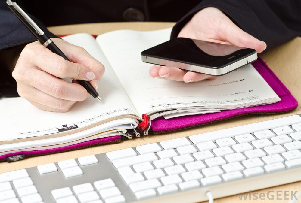 A person writing on a notebook near a keyboard while holding a cellphone